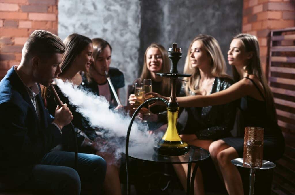 Friends party in hookah lounge. Group of people women and men smoking shisha in cafe or bar, making smoke clouds, having fun, smiling. Relax concept. Friendship